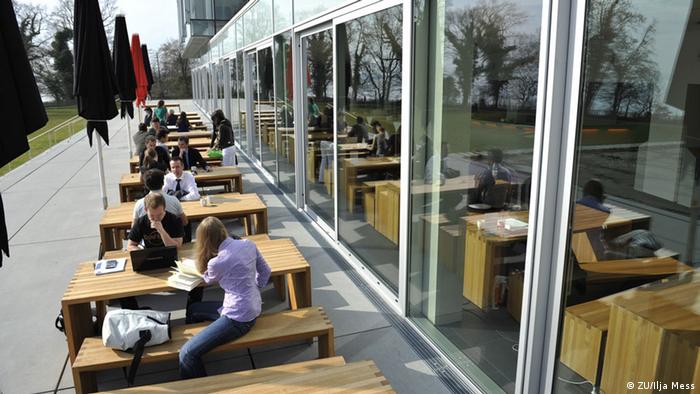 Na fotografiji su prikazani studenti sveučilišta Zeppelin koji sjede za drvenim stolovima izvan zgrade sveučilišta i čitaju iz knjiga ili sa svojih laptopa. 